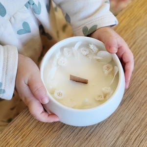 Peut inclure: Une bougie en céramique blanche avec une mèche en bois, ornée de fleurs séchées. La bougie est tenue par une personne portant une combinaison blanche à motifs de cœurs bleus. La bougie repose sur une surface en bois.