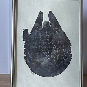 Peut inclure: Une impression en noir et blanc du vaisseau spatial Millennium Falcon dans un cadre argenté. L'impression a un fond texturé et moucheté.