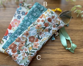 May include: Three fabric eyeglass cases with floral patterns. The cases are in shades of white, teal, and cream, with orange, yellow, and green floral designs. One case has a pair of sunglasses inside, with a green ribbon attached.