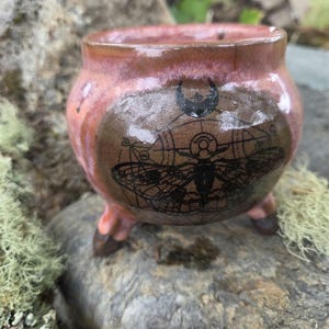 Può includere: Un piccolo calderone in ceramica a tre gambe, di forma rotonda, con una smaltatura lucida rosa e marrone. La parte anteriore presenta una stampa grafica nera, scura e dettagliata, di una falena con linee geometriche e una luna.