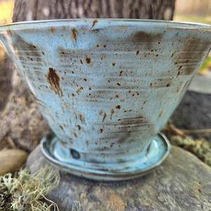 May include: A light blue ceramic planter with a wide, flared rim and a matching circular saucer base. The exterior features a mottled glaze with dark brown speckles and horizontal ridges, creating a textured appearance across the surface.