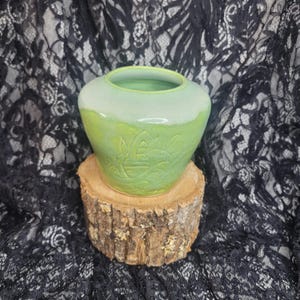May include: A green ceramic vase with a carved floral design, on a small wooden stand. The vase has a rounded shape and a light green top, with a darker green base. The background is a black lace fabric.