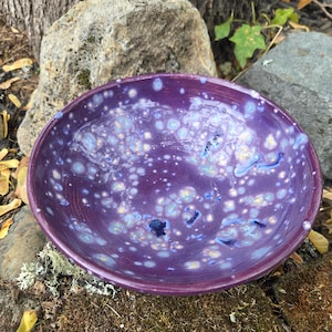 May include: A round ceramic bowl with a glossy purple glaze featuring a speckled pattern of white, light blue, and dark blue spots. The interior surface has a smooth, reflective finish with visible variations in the glaze texture and color density.