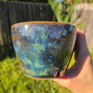May include: A ceramic bowl featuring a glossy glaze with a blend of deep blue, vibrant turquoise, and earthy brown tones. The surface exhibits a mottled, variegated appearance with smooth, rounded edges and a wide, open circular rim design.