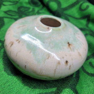 Op de afbeelding: Kleine, ronde keramische vaas met een brede body en een kleine opening. Crèmewitte basis met strepen van lichtgroen en bruine spikkels. De glanzende afwerking reflecteert licht, wat het gladde oppervlak benadrukt. Decoratief item.