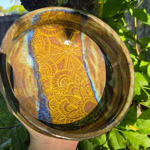 May include: A handmade ceramic bowl with a detailed yellow mandala design on a brown background. The bowl has a dark brown rim and blue accents. The bowl is held outdoors with green foliage in the background.