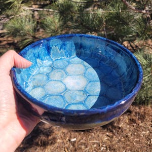 May include: A handmade ceramic bowl with a glossy, deep blue glaze. The interior features a honeycomb pattern in shades of blue and white. The bowl is held in a hand, showcasing its size and shape, perfect for serving or display.