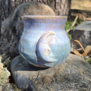 May include: A rounded ceramic mug featuring a glossy, variegated glaze in shades of periwinkle blue, lavender, and soft gray. A raised, sculpted crescent moon with a detailed facial profile is embossed on the front surface of the vessel.