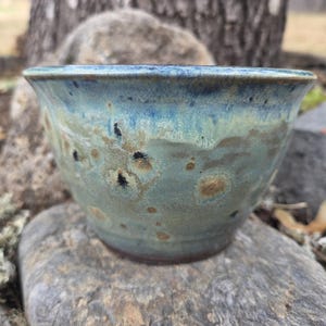 May include: A ceramic bowl featuring a variegated glaze with blended tones of deep blue, sage green, and earthy brown. The surface displays a mottled texture with dark speckles and lighter circular patterns, finished with a smooth, flared rim.
