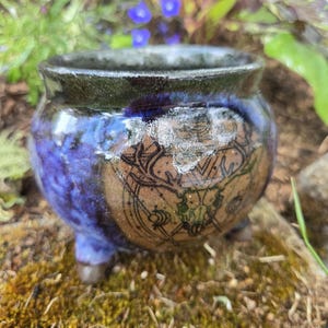 Può includere: Un piccolo calderone in ceramica a tre gambe con finitura lucida. L'esterno presenta uno smalto blu profondo e nero, con un pannello circolare centrale color marrone chiaro che mostra un'illustrazione a linee nere di un teschio di cervo.