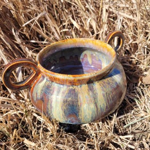 May include: A handmade ceramic bowl with two loop handles. The bowl features a glossy glaze with a blend of colors, including brown, gold, blue, and green. The bowl is sitting on a bed of dry grass.