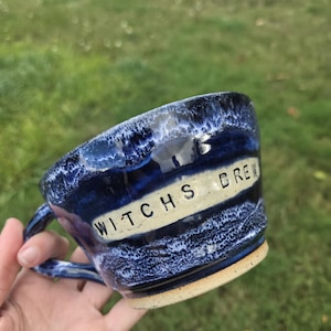 May include: A ceramic mug with a deep blue and white mottled glaze featuring a horizontal cream-colored band across the center. The text WITCHS BREW is stamped in black capital letters on the band. The mug has a sturdy handle and a base.
