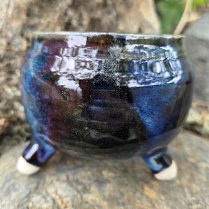 Puede incluir: Un pequeño recipiente de cerámica redondo con un esmalte brillante en tonos azul oscuro y negro. La pieza presenta una abertura ancha y se apoya sobre tres patas cortas y robustas con puntas redondeadas de color claro, creando una base estable.