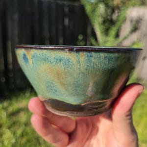 May include: A small ceramic bowl featuring a glossy glaze with blended shades of teal, green, and brown. The rim is dark and defined, while the lower portion of the bowl transitions into a solid dark brown base with a smooth, rounded shape.