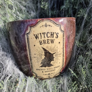 May include: A ceramic mug with a glossy, mottled brown and purple glaze. The front features a cream-colored label with the black text Witch's Brew, a silhouette of a witch in a pointed hat, and the text Brewed with Magic & Mischief.