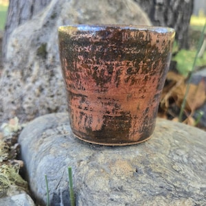 May include: A small, cylindrical ceramic cup featuring a mottled, rustic glaze. The surface displays a blend of earthy terracotta orange and deep, dark brown tones with a textured, uneven finish that creates a weathered, organic appearance.