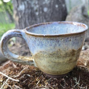 Könnte beinhalten: Eine handgefertigte Keramik-Tasse mit abgerundeter Form und einem stabilen Henkel. Die Tasse weist einen Farbverlauf auf, der von einem hellen Blau oben zu einem warmen, erdigen Braun am Boden übergeht. Die Tasse ist zum Trinken von Getränken geeignet.