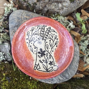 May include: A round ceramic dish with a glossy orange-red rim and a cream-colored center. The center features a black line-art illustration of a rabbit surrounded by stylized floral and leaf patterns, with an eye symbol on the rabbit's head.