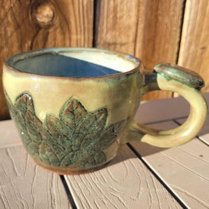 May include: Handmade ceramic mug with a leaf design. The mug has a light green exterior with brown and green leaf imprints. The interior is a light blue colour. The handle has a leaf-shaped accent. The mug is on a white surface.
