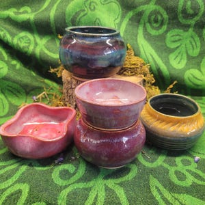 Puede incluir: Una colección de cuencos de cerámica hechos a mano en varias formas y tamaños. Los cuencos presentan una gama de colores, incluyendo azules profundos, morados y amarillos. Algunos cuencos tienen superficies texturizadas, mientras que otros son lisos. Los cuencos se exhiben sobre una tela verde estampada.