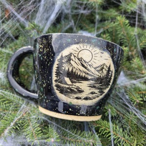 May include: A dark blue ceramic mug with a speckled finish and a large, circular cream-colored panel featuring a black ink illustration of a mountain range, a forest, a river, and a small tent under a sun with radiating rays.