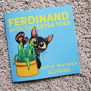 May include: A blue book cover with the title "Ferdinand with the Extra Toes" in yellow letters. The cover features a black cat with large eyes peeking out from behind a green plant in a yellow pot. The author is Katie Maskey with illustrations by Ashley Thoreen.