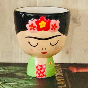 May include: A ceramic planter in the shape of a face with closed eyes, black hair, and a floral crown. The planter has a light skin tone, a green top, and a red and white patterned design on the front. The planter is on a dark surface.