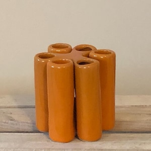 May include: A ceramic vase with seven cylindrical openings, arranged in a circular pattern. The vase is a vibrant orange colour and sits on a wooden surface. The design is modern and decorative.