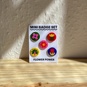 May include: A white rectangular card labelled MINI BADGE SET and FLOWER POWER featuring five circular pin-back buttons. The buttons display colourful floral illustrations in green, red, pink, yellow, and blue with various flower shapes.