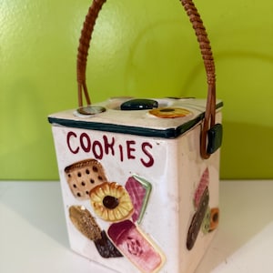Peut inclure: Un pot à biscuits en céramique vintage avec une poignée tressée. Le pot blanc porte l'inscription "COOKIES" en rouge et est décoré d'images colorées en relief de divers biscuits. Le couvercle comporte des éléments décoratifs.