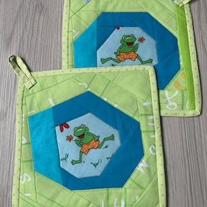 Könnte beinhalten: Zwei quadratische, gesteppte Topflappen. Jeder Topflappen zeigt einen Cartoon-Frosch in orangefarbenen Shorts, der eine Blume hält, vor blauem Hintergrund. Die Topflappen haben ein geometrisches Design in Grün und Blau. Mit einer Schlaufe zum Aufhängen.
