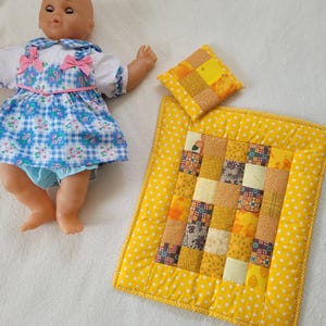 Könnte beinhalten: Eine Puppe in einem blau-weiß karierten Kleid mit rosa Schleifen, neben einer gelben Patchwork-Quilt und einem passenden Kissen. Der Quilt hat einen weißen Sternmuster-Rand.