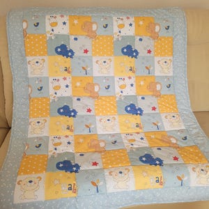 Könnte beinhalten: Eine Patchwork-Babydecke mit hellblauem Rand. Die Decke zeigt Quadrate mit Illustrationen von Bären, Elefanten, Wolken, Sternen und dem Wort "Baby". Die Quadrate sind in Gelb-, Blau- und Weißtönen gehalten.