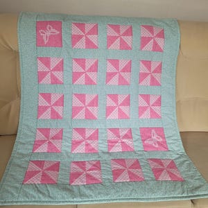 Könnte beinhalten: Handgefertigte Steppdecke mit einem Windrad-Design in Rosa und Hellblau. Die Steppdecke hat einen hellblauen Rand und zwei Schmetterlingsapplikationen. Die Windradblöcke sind in einem Raster angeordnet.