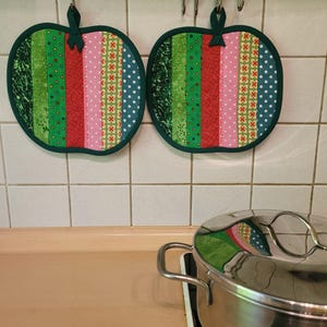 Könnte beinhalten: Zwei apfelförmige Topflappen aus grünem, rotem, rosa und blau gemustertem Stoff. Sie hängen an einer Metallstange vor einer weiß gefliesten Wand. Ein Edelstahltopf steht auf einem Herd.