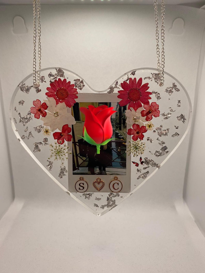 May include: A heart-shaped resin ornament with a silver chain, featuring a red rose, dried flowers, and silver leaf accents. The ornament also includes a photo with the letters "S", a heart, and "C" hanging from it.