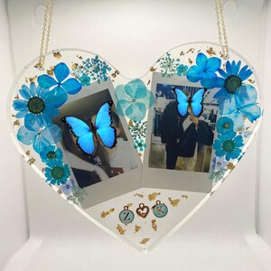 May include: A heart-shaped resin picture frame with two photo slots, decorated with pressed blue flowers, gold leaf, and butterflies. The frame is hanging from a gold chain and has two small charms with the letters "Z" and "K" on them.