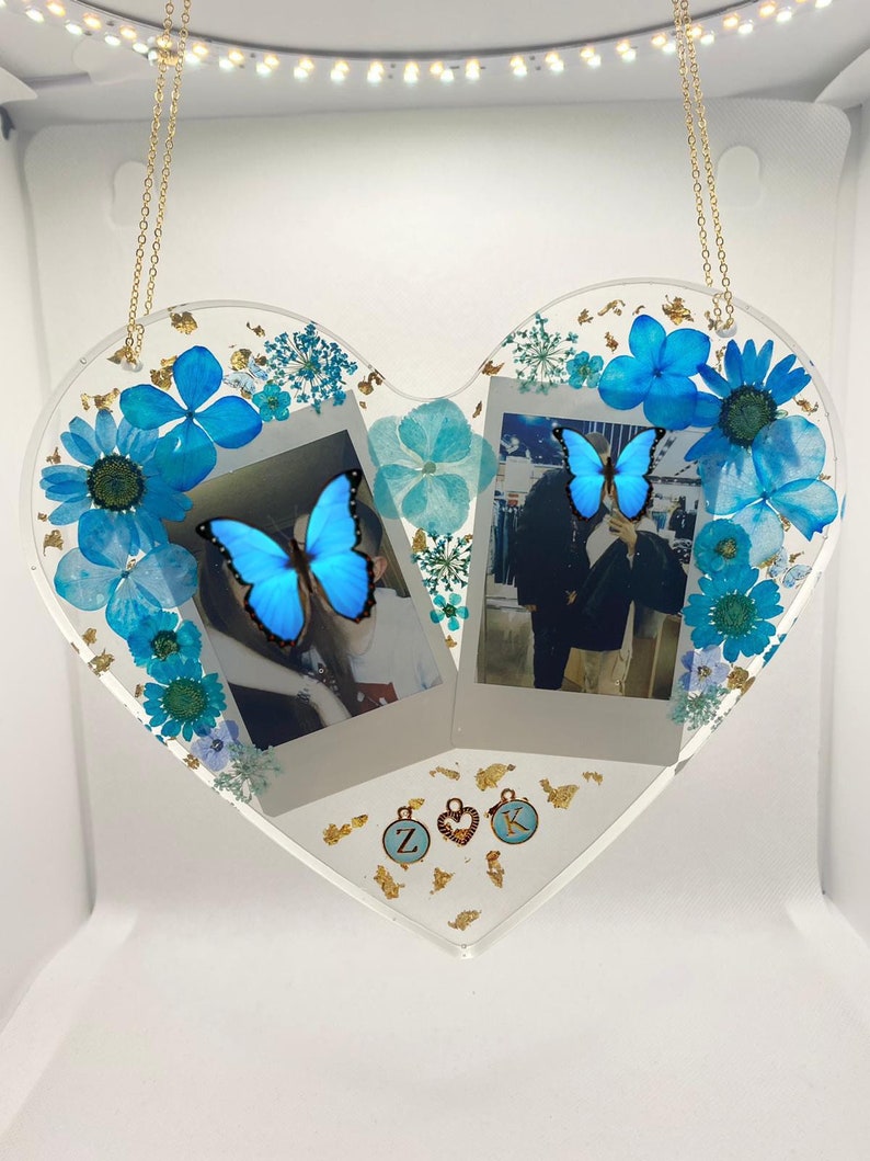 May include: A heart-shaped resin picture frame with two photo slots, decorated with pressed blue flowers, gold leaf, and butterflies. The frame is hanging from a gold chain and has two small charms with the letters "Z" and "K" on them.