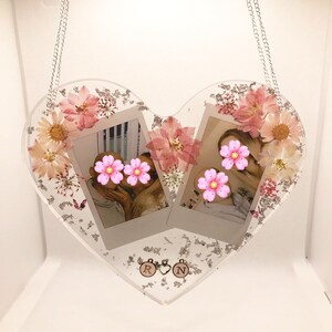 May include: A heart-shaped resin picture frame with two photo slots, decorated with dried flowers and glitter. The frame is hanging from a silver chain and has two charms with the letters "R" and "N".
