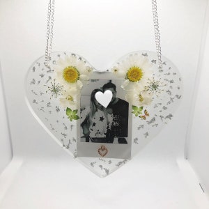 May include: A clear heart-shaped resin ornament with a silver glitter background. The ornament features a black and white photo of a couple, a small white heart over their faces, and a small gold heart charm hanging from the bottom. The ornament is decorated with white daisies and a butterfly.