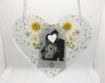 May include: A clear heart-shaped resin ornament with a silver glitter background. The ornament features a black and white photo of a couple, a small white heart over their faces, and a small gold heart charm hanging from the bottom. The ornament is decorated with white daisies and a butterfly.