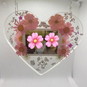 May include: A heart-shaped resin picture frame with a photo of two people and pressed flowers. The frame is hanging from a silver chain and has a silver heart charm.