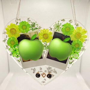 May include: A clear heart-shaped resin picture frame with two photo slots, featuring green pressed flowers and leaves, two green apples, and gold charms with the letter "C" and a heart.