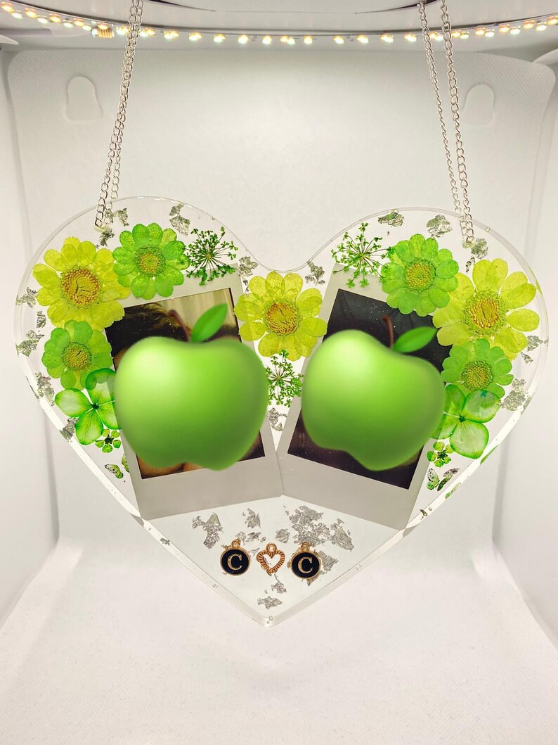 May include: A clear heart-shaped resin picture frame with two photo slots, featuring green pressed flowers and leaves, two green apples, and gold charms with the letter "C" and a heart.
