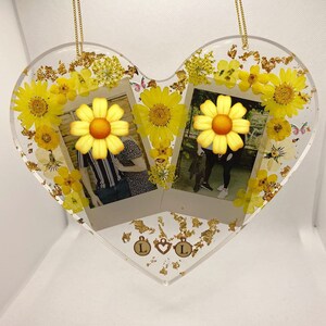 May include: A heart-shaped resin ornament with two Polaroid photos of a couple, surrounded by pressed yellow flowers and gold leaf. The ornament has two gold charms with the letter "L" on them.