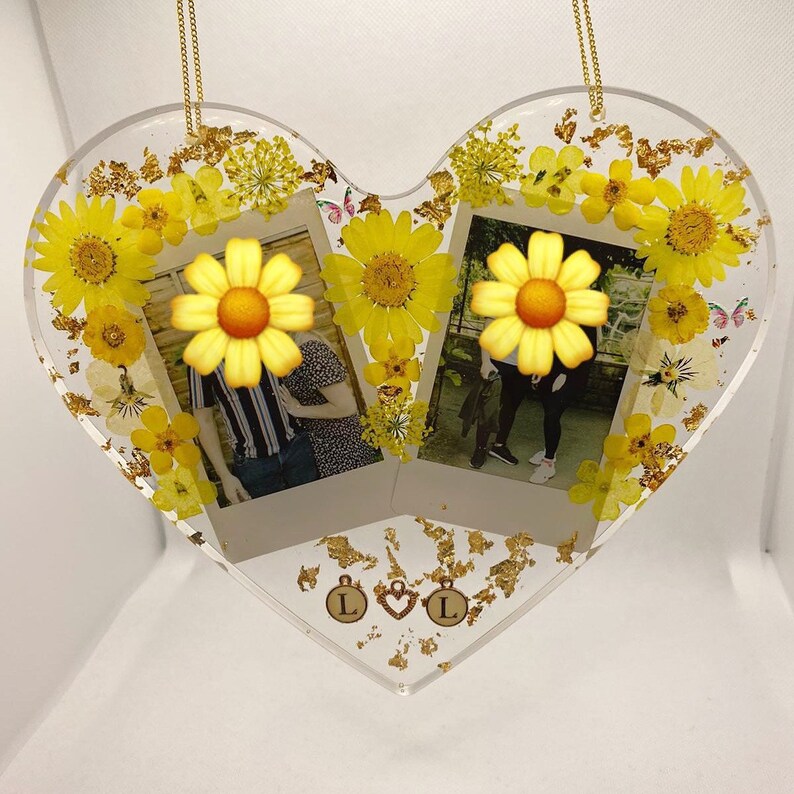 May include: A heart-shaped resin ornament with two Polaroid photos of a couple, surrounded by pressed yellow flowers and gold leaf. The ornament has two gold charms with the letter "L" on them.