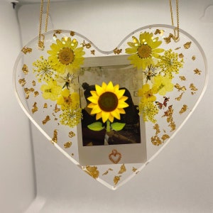 May include: A heart-shaped resin ornament with a pressed sunflower, dried flowers, and gold leaf accents. A small photo is embedded in the center of the ornament.