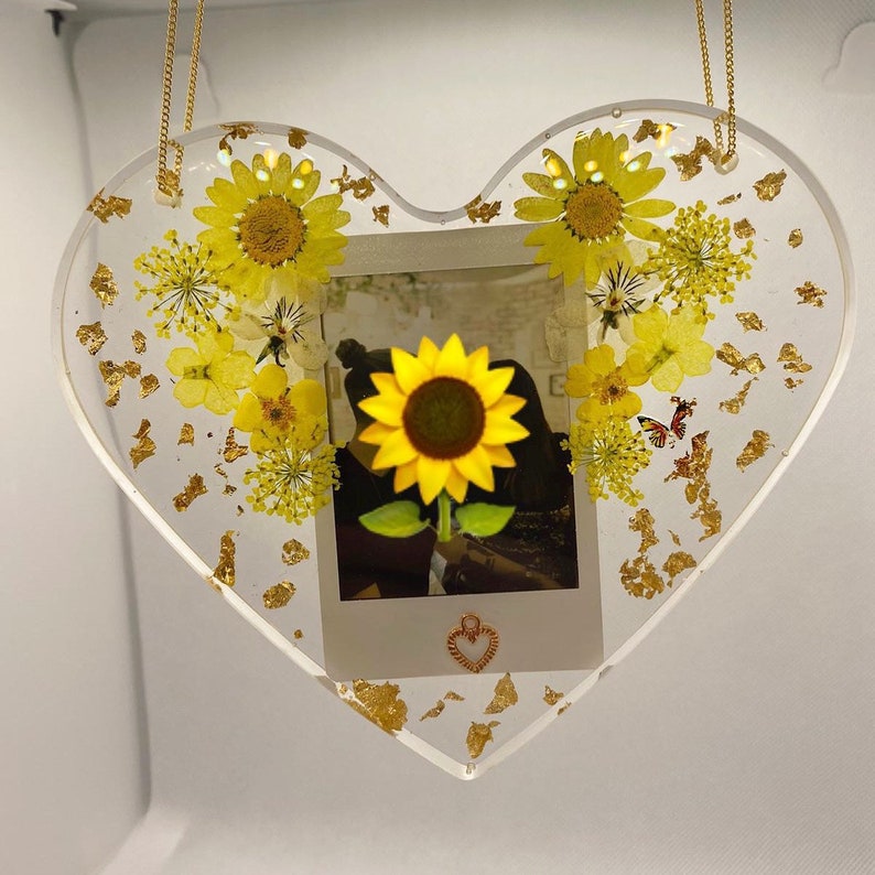 May include: A heart-shaped resin ornament with a pressed sunflower, dried flowers, and gold leaf accents. A small photo is embedded in the center of the ornament.