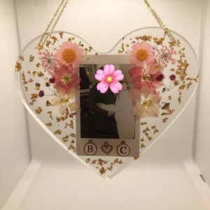 May include: A heart-shaped resin ornament with a photo of a couple, pink pressed flowers, and gold leaf accents. The ornament has a gold chain for hanging and the letters "B" and "C" are visible at the bottom.