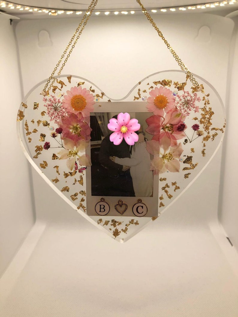 May include: A heart-shaped resin ornament with a photo of a couple, pink pressed flowers, and gold leaf accents. The ornament has a gold chain for hanging and the letters "B" and "C" are visible at the bottom.